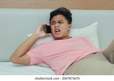 Portrait of unpleasant Asian man in pink t-shirt getting angry during phone conversation while lying on bed in the bedroom - Powered by Shutterstock