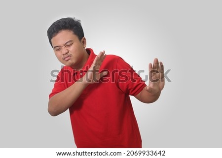 Portrait of unpleasant Asian man in red polo shirt forming a hand gesture to avoid something. Advertising concept. Isolated image on gray background