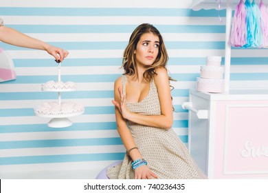 Portrait Of Unpleased Young Woman Turn Away From Tasty Cakes Posing On Cute Striped Background. Elegant Girl In Trendy Dress And Blue Accessories Refuse To Eat Sweet Dessert And Standing With Hand Up.