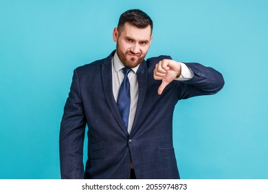 Portrait of unsatisfied bearded man wearing official style suit showing thumb down and looking at camera, dislike, application testing feedback. Indoor studio shot isolated on blue background.
