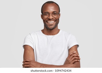 Portrait Of Unshaven Pleased Dark Skinned Male Has Warm Broad Smile, Keeps Hands Crossed, Rejoices Hearing Positive News, Wears Casual White T Shirt And Glasses. People And Positive Emotions