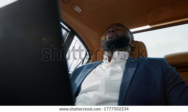 Portrait of upset afro man getting bad news at\
luxury car. Sad african businessman using laptop computer in\
vehicle. Disappointed professional feeling upset of project results\
at remote workplace
