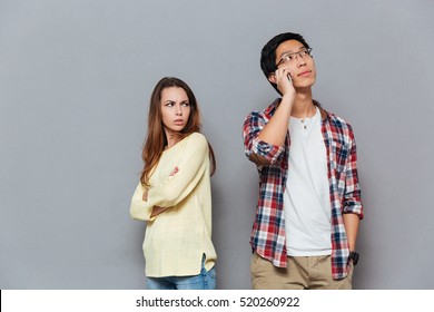 Portrait of upset angry girl watching her boyfriend talking on mobile phone isolated on the gray background