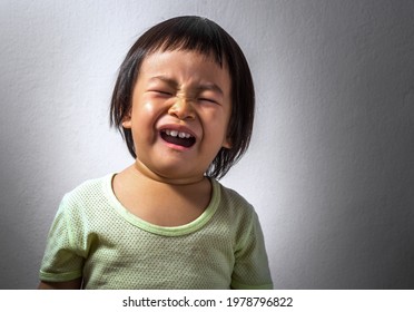 Portrait Of An Upset Asian Toddler Girl, Crying Alone, The Background Of Dark White Wall, Blank Space For Text.