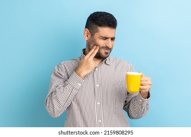Portrait of upset bearded businessman suffering from terrible teeth pain after drinking hot or cold beverage, dental injury, wearing striped shirt. Indoor studio shot isolated on blue background.