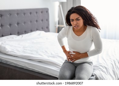 Portrait Of Upset Black Woman Suffering From Strong Abdominal Pain In Right Side, Touching Her Tummy, Sitting In Bed At Home. Sad Afro Lady Feeling Acute Stomachache, Free Copy Space