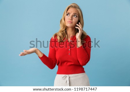 Portrait of upset blond woman 20s wearing red shirt talking on smartphone isolated over blue background in studio