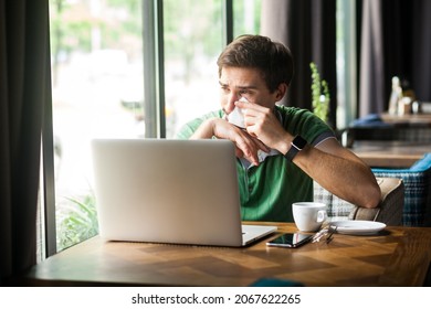 Portrait of upset businessman wearing green T-shirt, sitting in front laptop, crying and cleaning his tears with tissue, expressing sadness. Indoor shot near big window, cafe background.
