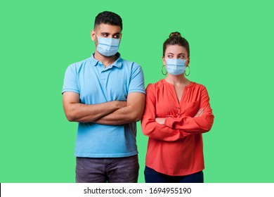 Portrait of upset couple with surgical medical mask standing together with crossed hands, looking sideways at each other with resentful glance, suspicion. isolated on green background, indoor studio