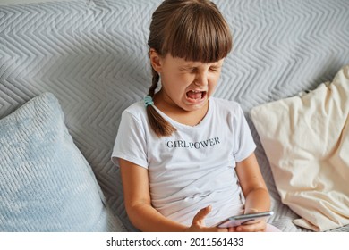 Portrait Of Upset Cute Crying Child With Dark Hair And Pigtails, Sitting On Sofa With Cell Phone In Hands, Being Sad Of Loosing Level In Favorite Game, Wearing White T Shirt.