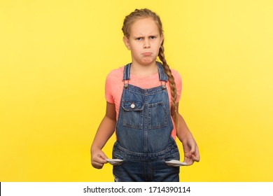 Portrait of upset little girl with braid in denim overalls turning out empty pockets, having no money, looking with depressed vexed expression. indoor studio shot isolated on yellow background