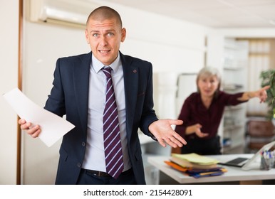 Portrait Of Upset Perplexed Office Employee Standing With Papers In Hands On Background Of Disgruntled Female Boss Reprimanding Him
