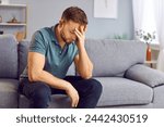 Portrait of upset sad depressed man sitting on sofa in living room at home alone suffering from headache. Young guy in bad thoughts feeling lonely indoors or having problems in relationships