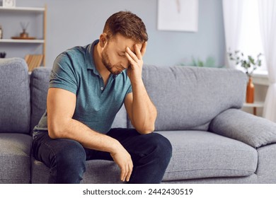 Portrait of upset sad depressed man sitting on sofa in living room at home alone suffering from headache. Young guy in bad thoughts feeling lonely indoors or having problems in relationships - Powered by Shutterstock