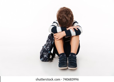 Portrait Of An Upset Sad Little Kid With Backpack Sitting On A Flor And Crying Isolated Over White Background