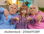 Portrait of upset senior grandparents man woman and granddaughter showing thumbs down gesture expressing discontent disapproval dissatisfied. Displeased family looking at camera on sofa in home room