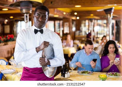 Portrait Of Upset Tired Adult African American Waiter Standing With Empty Serving Tray In Busy Restaurant Hall..