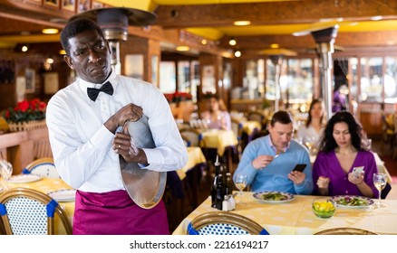 Portrait Of Upset Tired Adult African American Waiter Standing With Empty Serving Tray In Busy Restaurant Hall..