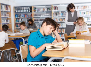 Portrait Of Upset Tween Boy Reading In School Library On Background With Other Students And Teacher