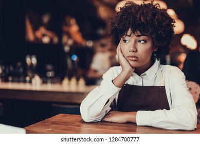 Portrait of upset waitress. She looking aside and sitting behind table while touching cheek by palm hand