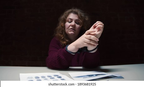 Portrait of upset woman criminal in handcuffs sitting at table with evidence in interrogation room. Suspect female handcuffed sitting in police department waiting for detective