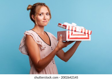 Portrait Of Upset Young Adult Blonde Female With Bundles, Holding Red Striped Present Box, Has Sad Expression, Being Disappointed With Birthday Gift. Indoor Studio Shot Isolated On Blue Background.