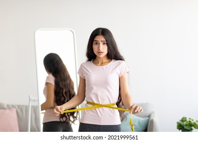 Portrait of upset young Indian lady measuring her waist with tape near mirror, feeling disappointed with results of slimming diet, unhappy about her body parameters at home