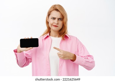 Portrait Of Upset Young Woman, Pointing At Her Mobile Phone With Frowned Angry Face, Stands Over White Background