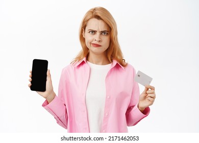 Portrait Of Upset Young Woman Showing Smartphone Application, Screen Of Mobile Phone And Credit Card, Frowning Angry, Standing Over White Background