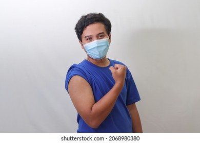 Portrait Of Vaccinated Asian Man In Blue T-shirt Wearing Medical Mask, Showing Strong Arm Muscle After Getting Vaccination. Isolated Image On Gray Background