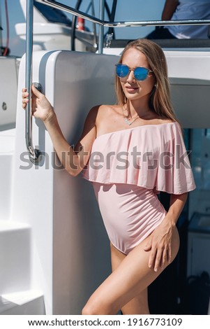 Portrait of a very beautiful girl in a tight pink bodysuit with ruffles and glasses posing on a white yacht, holding on to the handrail. Summer concept.