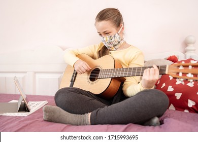 Portrait of very cute teenager in antibacterial mask playing her guitar sitting on the bed at home due to the isolation of COVID-19. Musician kid is using her tablet for online music lesson.