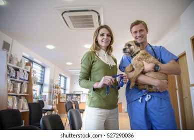 Portrait Of Vet Holding Dog With Female Owner In Veterinary Waiting Room