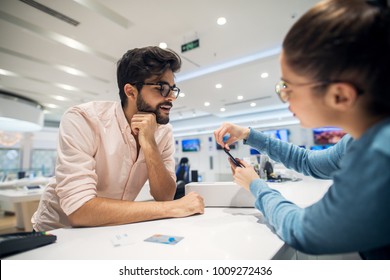 Portrait View Of Curious Excited Happy Smiling Young Student Bearded Man Looking At Mobile While Female Seller Giving Instruction For Sim Place On The Phone After Buying With Bank Card In A Tech Store