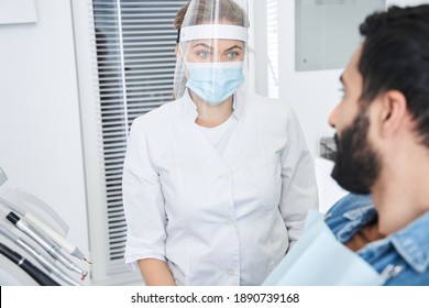 Portrait View Of The Female Dentist Wearing Protective Mask Talking With Her Male Patient And Preparing For Treatment In Dental Office. Bearded Multiracial Man In Dentist Chair Looking At His Doctor