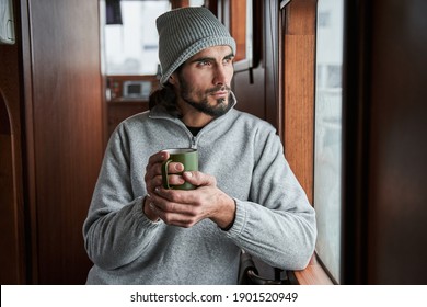 Portrait view of the handsome man wearing warm clothes is smiling and drinking coffee in the morning at the boat before the work. Fisherman warming with hot beverage before the fishing at the winter