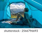 Portrait of a view from tent entrance of young traveler man relaxing in mountains. Rear view of hiker male sitting in the camping tent.