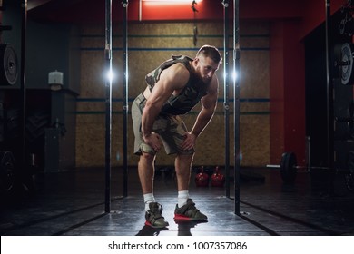 Portrait view of young bearded focused strong muscular shape bodybuilder man in military vest crouching with hands on knees and taking a break after exhausting training at the gym. - Powered by Shutterstock