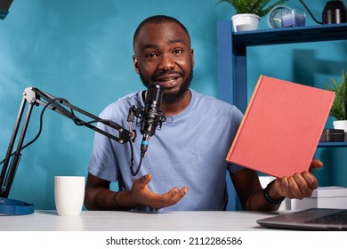 Portrait Of Vlogger With Audio Microphone Recording Podcast Talking About The Story In Red Mockup Book In Vlogging Studio. Content Creator Sitting At Desk Holding New Novel For Social Media Video.