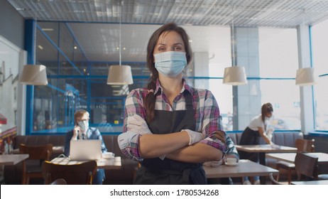 Portrait Of Waitress Wearing Face Mask And Gloves While Serving In Coffee Shop. Pretty Restaurant Worker In Apron And Protective Mask And Gloves Posing At Camera With Crossed Hands