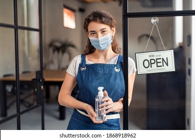 Portrait Of Waitress Wearing Face Mask And Holding A Bottle Of Sanitiser At Cafe Entrance During Covid-19 Pandemic. Small Business Owner Holding Sanitizer Reopening After Coronavirus Lockdown.