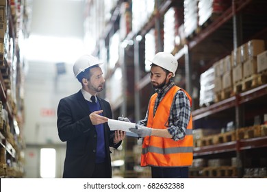 Portrait Of Warehouse Manager Wearing Business Suit Talking To Loader Showing Order List In Aisle Between Tall Shelves With Packed Goods