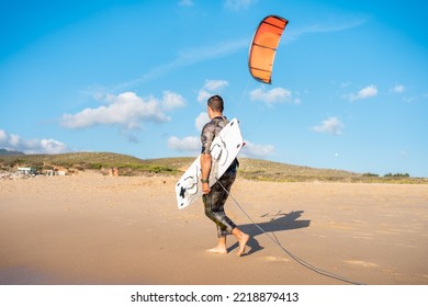 Portrait wave kitesurfer walking upwind at beach with his board and kite. Man kite surfer walk sand ocean beach with his kite surf board.