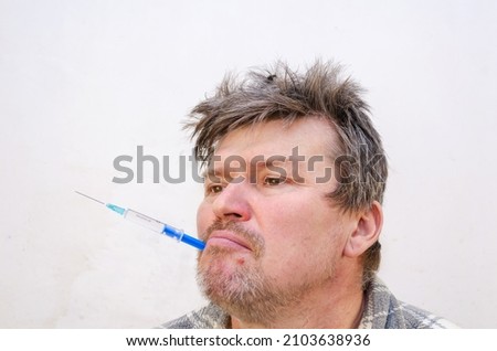 Portrait of a weirdo holding a blue syringe in his mouth. Unshaven man holding a disposable syringe full of liquid. Adult male with ruffled hair.  Indoors. Selective focus.