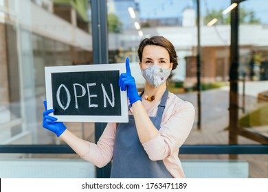 Portrait Of Welcoming Grocery Shop Female Owner Outdoor. Business Woman Worker In Apron Protective Mask And Gloves Holding Open Sign Placard At Entrance. Girl Open Door After Lockdown Quarantine.