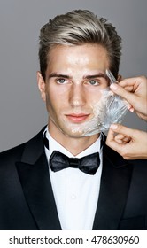 Portrait of well dressed elegant man in facial mask on gray background. Women's hands beautician removes peeling off a facial mask from the man's face. Grooming himself