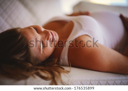A portrait of a white beautiful young pregnant woman lying on a white sofa and touching her baby bump with a happy smile of a future mother 