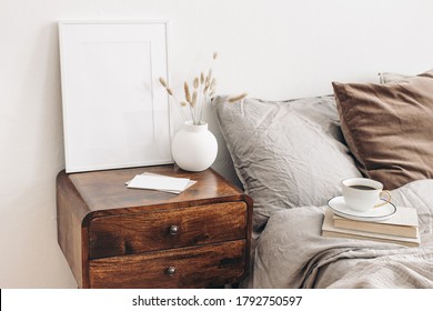 Portrait white frame mockup on retro wooden bedside table. Modern white ceramic vase, dry grass. Cup of coffee and books in bed. Beige linen pillows in bedroom. Scandinavian interior.