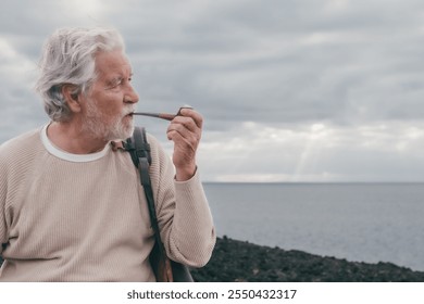 Portrait of white haired bearded senior man sitting along the rocky beach smoking pipe and looking away, horizon over sea - Powered by Shutterstock