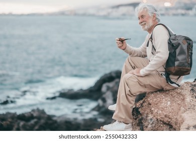 Portrait of white haired bearded senior man sitting along the rocky beach smoking pipe and looking away, horizon over sea - Powered by Shutterstock